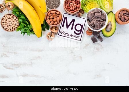 Lebensmittel, die natürliches Magnesium enthalten. Mg: Schokolade, Banane, Kakao, Nüsse, Avocados, Brokkoli, Mandeln. Draufsicht. Auf weißem Holzhintergrund. Stockfoto