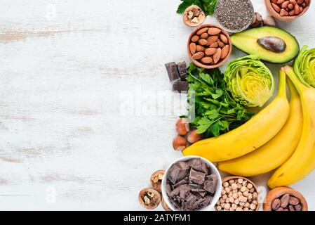 Lebensmittel, die natürliches Magnesium enthalten. Mg: Schokolade, Banane, Kakao, Nüsse, Avocados, Brokkoli, Mandeln. Draufsicht. Auf weißem Holzhintergrund. Stockfoto