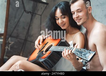 Gemischtes Rennpaar. Junger Mann, der Frau beigebracht hat, Gitarre auf dem Bett sitzend, lächelnd fröhlich zu spielen Stockfoto