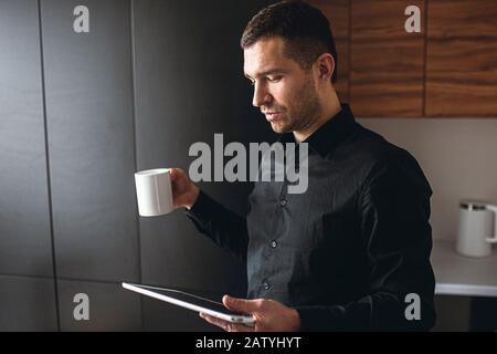 Seitenansicht des jungen Geschäftsmannes in der Küche arbeiten abgelegen. Unternehmer halten Tablette und sehen sich das an. Trinken Sie Tee während der Kaffeepause. Effizientes Arbeiten zu Hause. Stockfoto