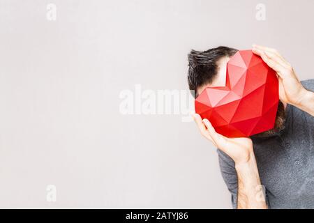 Junger Mann, der in der Ecke stehend polygonales rautenförmiges rotes Herz vor dem Kopf hält. Stockfoto