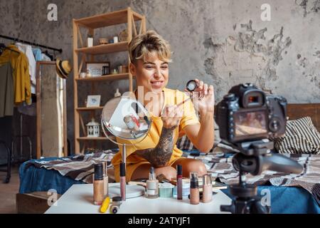 Make-up Blogger. Junge Frau, die in einer stilvollen Wohnung sitzt und Videos auf der Kamera aufzeichnet, die ein Produkt mit Augenweise zeigt Stockfoto
