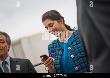 Lächelnde junge Geschäftsfrau checkt ihr Telefon bei einem Geschäftstreffen auf dem Balkon eines Hochhauses. Stockfoto
