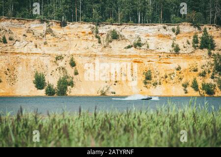 Hohe felsige sandige Steilufer mit Pinien bedeckt über einem klaren und blauen See. Schnelle Motorboot schneidet durch die Wasseroberfläche Stockfoto