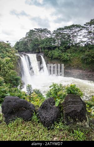 Hilo, Hawaii, USA. - 14. Januar 2020: White Rainbow Falls on Foaming Violent Wailuku River umgeben von grünen Bäumen und Pflanzen unter weiß-grauem Clou Stockfoto