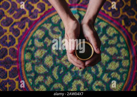 Ein Mann hält eine Tasse halluzinogenen Ayahuasca Pflanzenmedikaments in seinen ausgestreckten Händen über einen bestickten Shipibo-Festteppich. Stockfoto