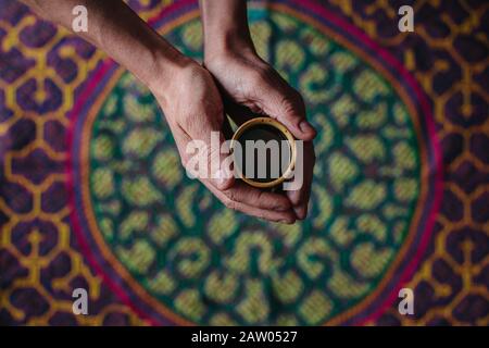 Ein Mann hält eine Tasse halluzinogenen Ayahuasca Pflanzenmedikaments in seinen ausgestreckten Händen über einen bestickten Shipibo-Festteppich. Stockfoto