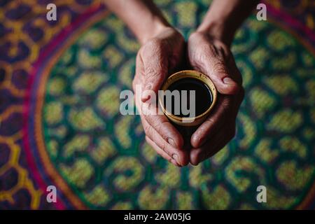 Ein Mann hält eine Tasse halluzinogenen Ayahuasca Pflanzenmedikaments in seinen ausgestreckten Händen über einen bestickten Shipibo-Festteppich. Stockfoto