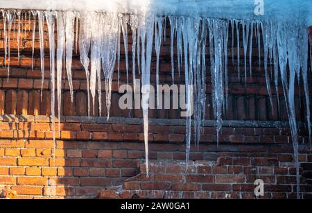 Große Eiszapfen hängen vom Dach eines alten Ziegelbaus. Dach des alten Gebäudes mit scharfen Eiszapfen bedeckt. Eis-Tropfstein. Schlechte Wärmedämmung Stockfoto