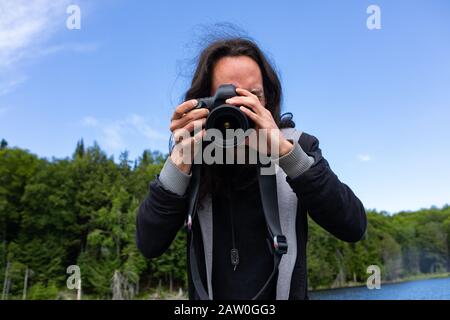 Junger amerikanischer Reisender mit langen Haaren fotografiert mit DSLR-Kamera, während er auf dem See mit Tannen und Fichten im Norden Quebecs, Kanada, reitet Stockfoto