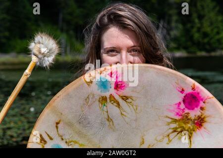 Nahaufnahme der schönen lächelnden Frau, die die heilige Rahmentrommel mit mit Pelz bedecktem Stock am See hält, mit Wasserlilien, die das Gesicht in Kanada bedecken Stockfoto