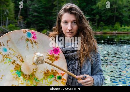 Porträt der jungen schamanischen Frau mit langen Haaren, die heilige einheimische Rahmentrommel mit mit Pelz überzogenen Stock in der Nähe von See mit Wasserlilien in Kanada spielt Stockfoto