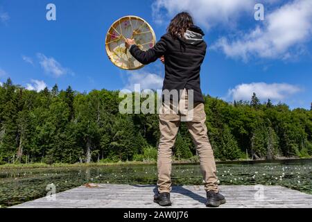 Lange Rückansicht des langhaarigen amerikanischen jungen Mannes, der auf dem Dock steht und die heilige einheimische Rahmentrommel spielt. Mit Pelz überdachter Stock in Kanada Stockfoto