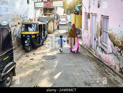 Udaipur, RAJASTHAN, INDIEN - 03. MÄRZ 2015: Frau in rosafarbenem Sari, die mit ihrem Sohn auf der schmalen Straße mit Rickshaw Taxi in Udaipur Cit zur Schule geht Stockfoto