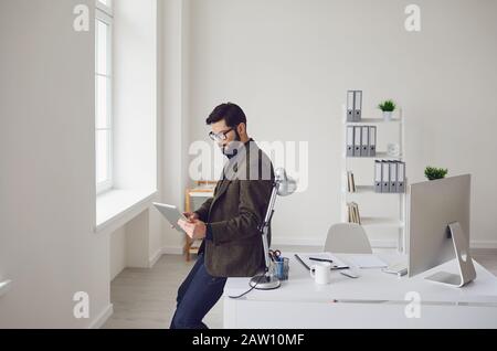 Der Geschäftsmann liest in einem Tablet, während er an einem Arbeitsplatz in einem weißen Büro steht. Stockfoto