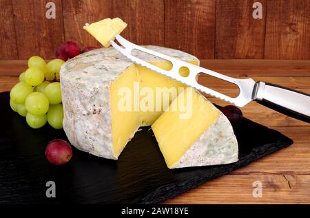 Käse aus Bauernhaus mit Käse-Messer und Käsekeil auf einer Schiefertafel mit Holzhintergrund Stockfoto