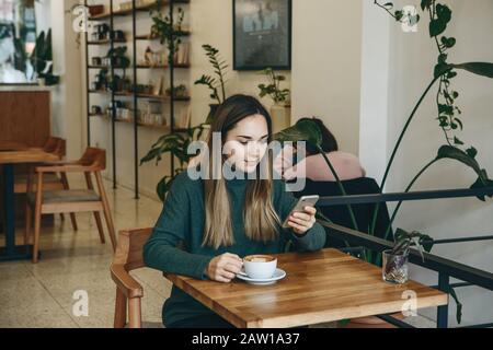 Das Mädchen verwendet ein Mobiltelefon und trinkt Kaffee. Sie hat ein Treffen oder sie trinkt einen morgendlichen Cappuccino oder flache weiße. Stockfoto