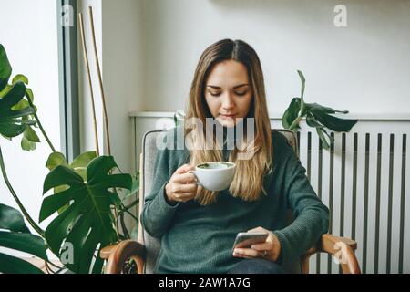 Mädchen trinkt ein warmes Morgengetränk und benutzt ein Handy. Stockfoto