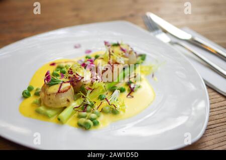 Gebratene Jakobsmuscheln mit gelber Sahnesauce, gestreutem Rotkohl, grünen Erbsen und violetten Blumen in weißer Platte Draufsicht Stockfoto