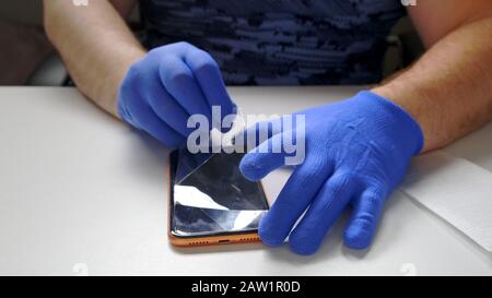 Die männliche Hand in blauen Handschuhen ersetzt einen zerbrochenen Schutzschirm aus gehärtetem Glas für ein Smartphone. Ein Mann bereitet ein Smartphone vor, um Glas zu ersetzen. Smartphone Stockfoto