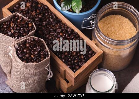 Jutebeutel, Holzbehälter mit Kaffeebohnen und Glasbecher mit Milch und braunem Sauger auf dem Hintergrund. Stockfoto