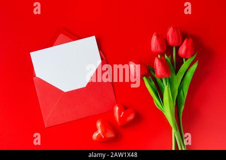 Grußkarte mit Liebe. Roter Umschlag mit leerem Papier, Blumenstrauß aus Tulpen und herzförmigen Kerzen. Draufsicht mit Platz für Ihren Text. Flaches Lay Stockfoto