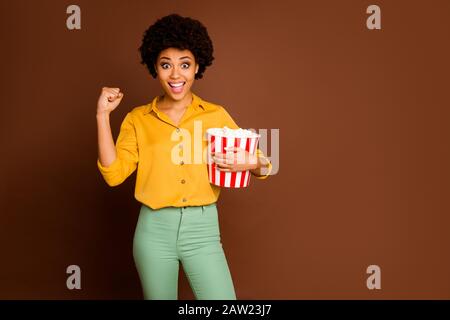 Foto der verrückten dunklen Haut gewellte Dame halten Popcorn Eat Corns beobachten Fußballspiel Unterstützung Lieblingsmannschaft tragen gelbes Hemd grüne Hosen isoliert braun Stockfoto