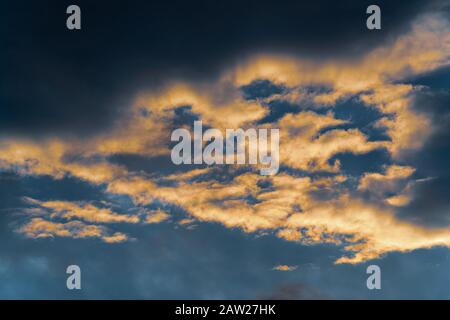 Goldene, flauschige Wolken, die durch verschwindende Strahlen bei Sonnenuntergang und dunkle Gewitterwolken erhellt werden, die über sonnigen blauen Himmel schweben, um das Wetter in der Saison zu ändern Stockfoto