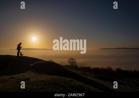 Ein Mann und sein Hund bewundern die Wolkeninversion über der Stadt Bath in Somerset bei Sonnenaufgang von Solsbury Hill an einem Wintertag. Stockfoto