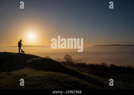 Ein Mann und sein Hund bewundern die Wolkeninversion über der Stadt Bath in Somerset bei Sonnenaufgang von Solsbury Hill an einem Wintertag. Stockfoto