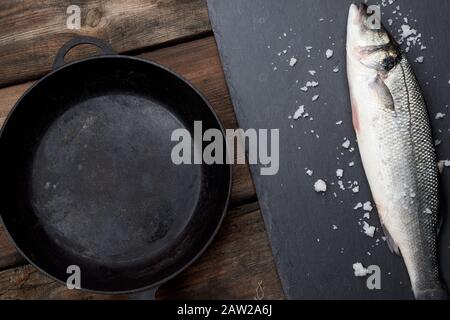 Frische ganze Wolfsbarsch Fisch auf einem schwarzen Brett, daneben ist eine leere runde Schwarze Pfanne auf einem Holztisch, Ansicht von oben Stockfoto