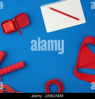 Rotes Sportshirt, rotes Springseil und kabellose Kopfhörer in roter Hülle auf blauem Grund, Draufsicht, leeres weißes Blatt Papier, flaches Lay Stockfoto