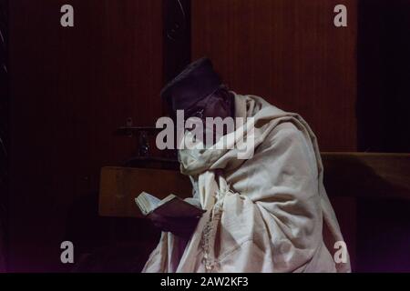 Lalibela, Äthiopien - Nov 2018: Mann kleidete sich in traditionelles Weiß, saß im Zwielicht und las vor den Untergrundkirchen von Lali ein Buch mit gebeten Stockfoto