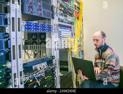 Ein Mann mit Laptop sitzt im Serverraum des Rechenzentrums. Der Systemadministrator arbeitet in der Nähe der Racks mit den Servern. Computeringenieur Stockfoto