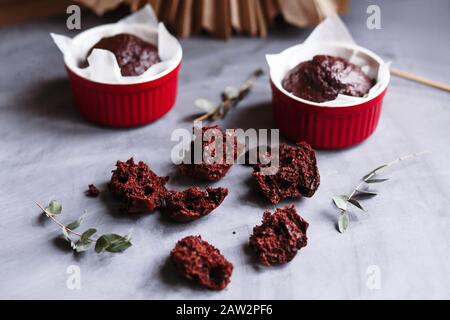 Schokoladenmuffins in roten Tassen. Kleines glasiertes Keramikramekin mit braunen Kuchen auf grauem und weißem Grund Stockfoto