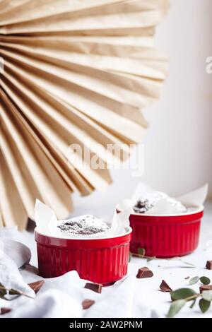 Schokoladenmuffins in roten Tassen. Kleines glasiertes Keramikramekin mit braunen Kuchen auf grauem und weißem Grund Stockfoto