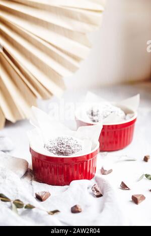 Schokoladenmuffins in roten Tassen. Kleines glasiertes Keramikramekin mit braunen Kuchen auf grauem und weißem Grund Stockfoto