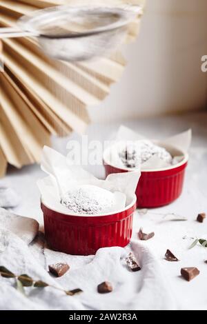 Schokoladenmuffins in roten Tassen. Kleines glasiertes Keramikramekin mit braunen Kuchen auf grauem und weißem Grund Stockfoto