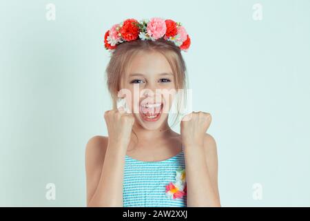 Fröhliches Mädchen übergibt sich jubelnd, feiernd, pumpt Fäuste, die glücklich auf die Kamera schauen. Nahaufnahme des kaukasischen Kindermodells mit geblümtem Stirnband Isol Stockfoto