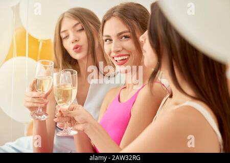 Ernte von drei jungen fröhlichen weiblichen Freunden, die feiern und Champagner in Gläsern im Schlafzimmer trinken. Selektiver Fokus der Gruppe von Mädchen lächelnd und mit Blick auf die Kamera. Festliche Stimmung, Ballons rund. Stockfoto