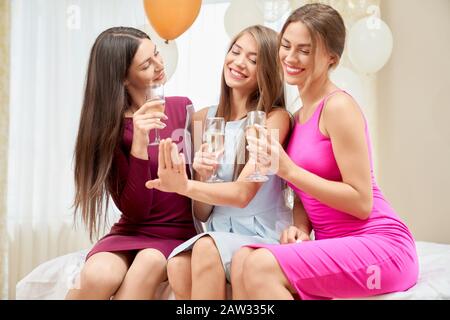 Ernte von drei jungen fröhlichen weiblichen Freunden, die feiern und Champagner in Gläsern im Schlafzimmer trinken. Selektiver Fokus der Gruppe von Mädchen lächeln und plaudern. Festliche Stimmung, Ballons rund. Stockfoto