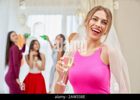 Vorderansicht der hübschen jungen lachenden Frau im Schleier, der morgens auf dem Bett sitzt und Glas mit Champagner hält. Schöne Frau, die sich auf der Junggesellen-Party lustig macht, Brautleute mit Flaschen hinterher. Stockfoto