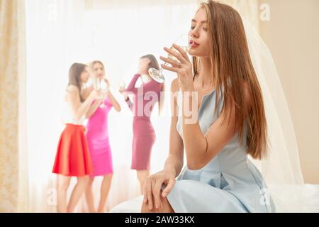 Blick auf die hübsche junge lächelnde Frau im Schleier, der morgens auf dem Bett sitzt und Champagner trinkt. Schöne Frau, die sich auf der Junggesellen-Party lustig macht, Brautleute mit Flaschen hinterher. Stockfoto