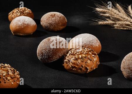 Auswahl an leckeren gebackenen Brötchen mit Sesam und Sonnenblumenkerne, bestreut mit Mehl, Haufen Weizen, vor schwarzem Hintergrund, Kopierraum. Ländliche Cuis Stockfoto