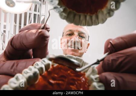 Gesicht des leitenden, konzentrierten männlichen Zahnarztes, der am Tisch sitzt und zahnmedizinische Werkzeuge in den Händen hält und Nachahmungsbehandlung am Zahnmodell der Kieferproben macht Stockfoto