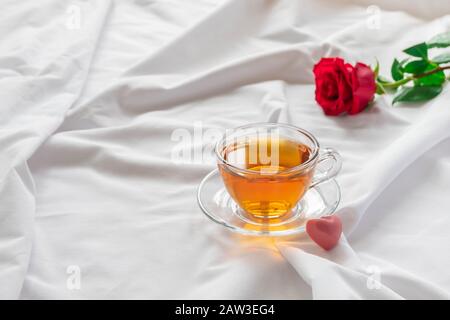 Romantisches Frühstück im Bett mit einer tasse Tee, herzförmiger rosafarbener Süßigkeiten und Rose auf weißen Bettlaken. Stockfoto
