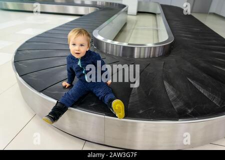 Allein niedlicher kaukasischer blonder Kleinkind, der auf dem leeren Gepäckförderband am Ankunftsterminal des Flughafens sitzt. Verlorenes gefundenes Kind. Mit dem Flugzeug reisen Stockfoto