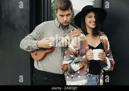 Stilvolles Hippie-Paar mit Morgenkaffee und lächelnder Ukulele, entspannend im Hintergrund der modernen Kabine mit großen Fenstern in den Bergen. Frohes junges Familienfest Stockfoto