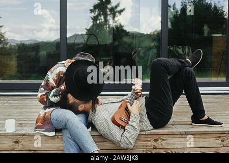 Der Mann von Hipper spielt auf Ukulele für seine schöne, stilvolle Frau, und entspannt sich auf der Holzterrasse im Hintergrund der modernen Kabine mit großen Fenstern in den Bergen. Stockfoto