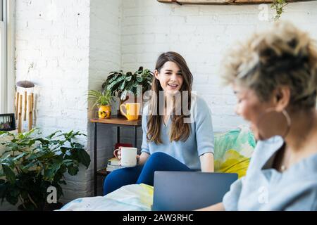 Lächelnde junge Frau, die mit einem Freund spricht, während sie zu Hause auf dem Bett sitzt Stockfoto